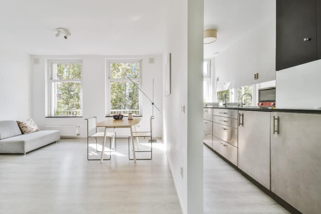 Intérieur moderne lumineux avec salon, salle à manger bois clair et cuisine design gris métal. Fenêtres sur arbres verts.