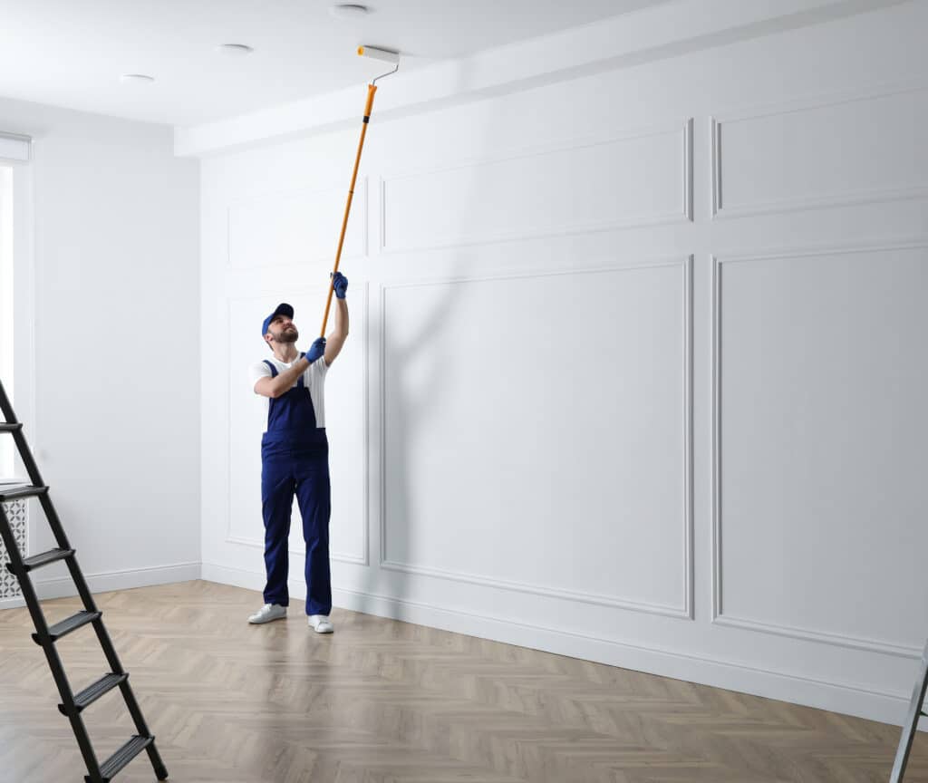 Homme peintre en salopette bleue peignant le plafond blanc d'une pièce avec un rouleau télescopique. Murs moulurés, sol en bois.