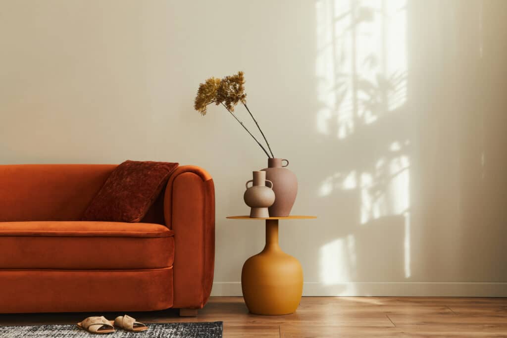 Salon moderne avec canapé orange velours, coussin marron, table d'appoint jaune, vases terre cuite, plante sèche sur mur beige clair et sol bois. Lumière du soleil.
