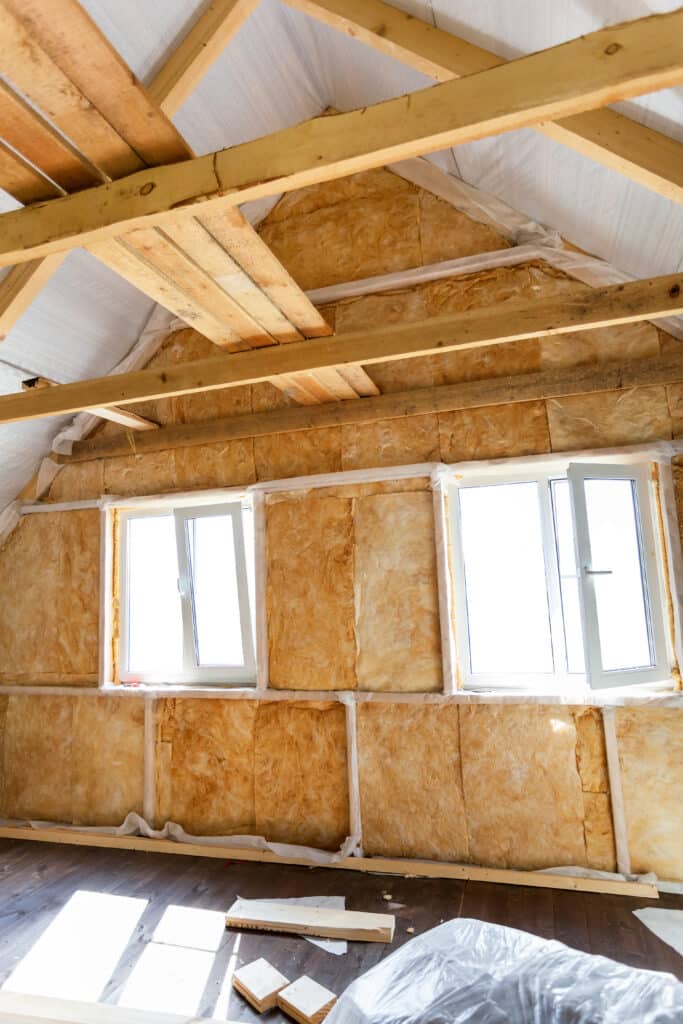 Intérieur de combles aménagés avec isolation en laine minérale, fenêtres PVC blanches, charpente bois apparente et plancher foncé éclairé.